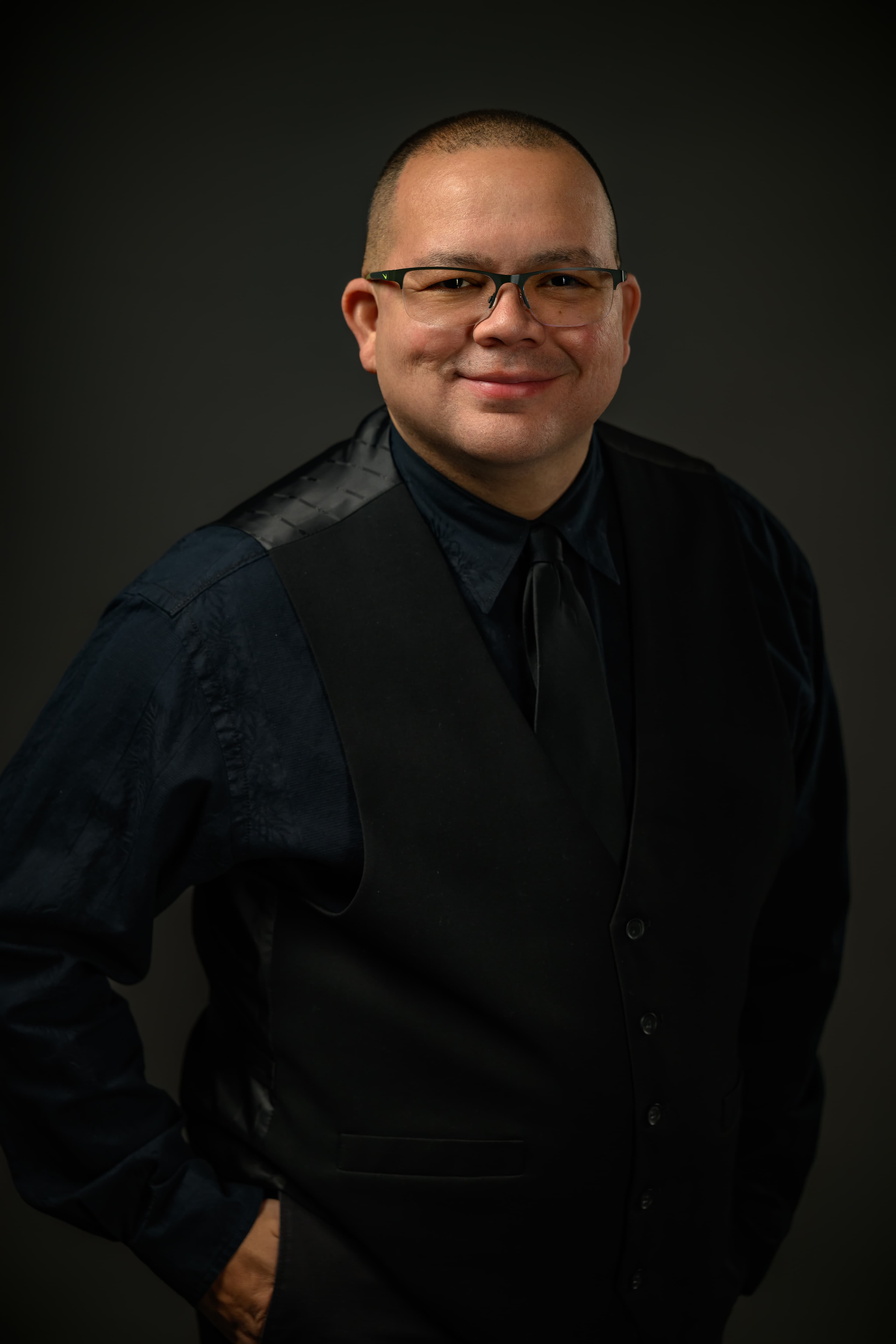 Smiling man with glasses wearing a black vest and tie, posed against a dark background.