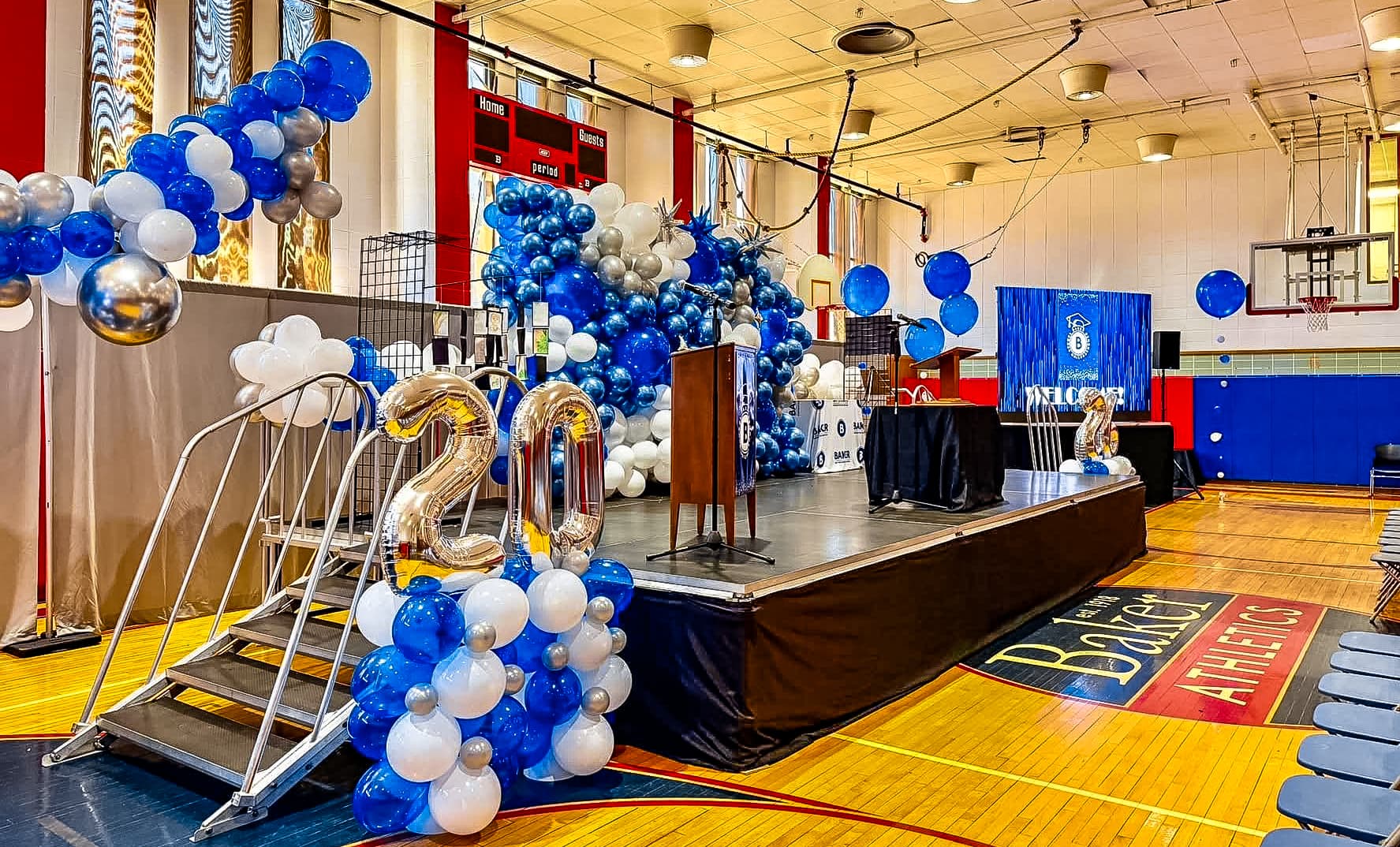 Gymnasium stage decorated with blue, white, and silver balloons and large gold "20" balloons.