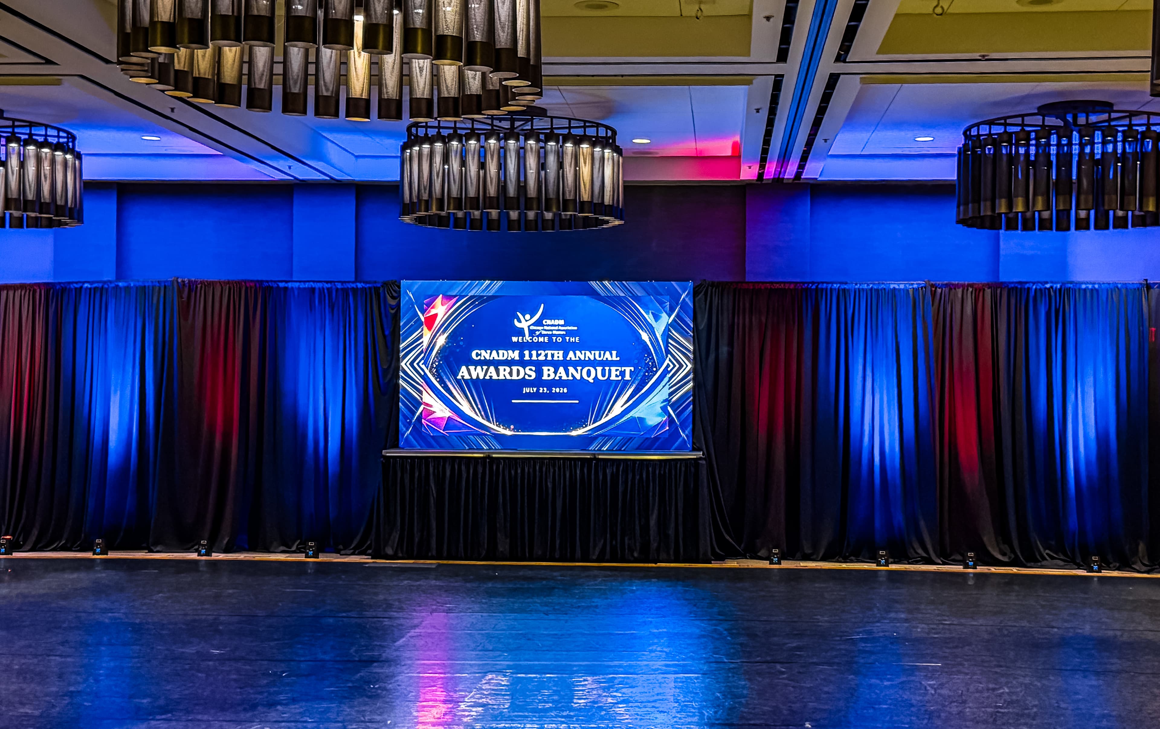 CNADM 112th Annual Awards Banquet stage featuring a large screen, blue lighting, and chandeliers.