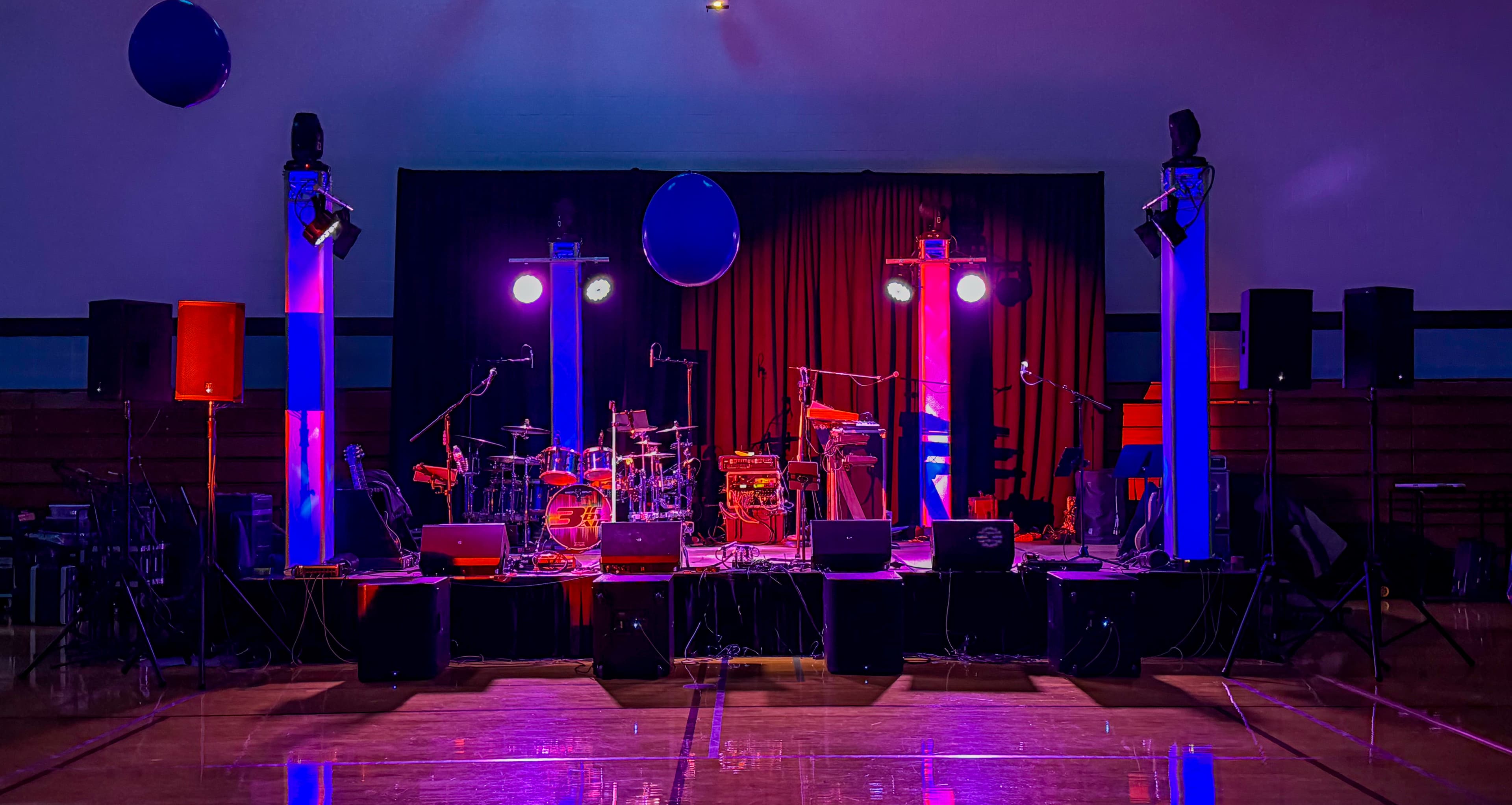 Stage with drums and speakers under dramatic blue and purple lighting in a gymnasium.
